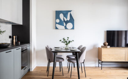 Nowoczesna jadalnia z czarnym stołem, czterema szarymi krzesłami i obrazem w odcieniach niebieskiego. W tle aneks kuchenny i drewniany stolik RTV.