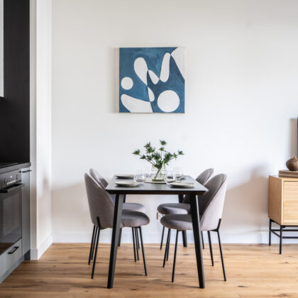 Nowoczesna jadalnia z czarnym stołem, czterema szarymi krzesłami i obrazem w odcieniach niebieskiego. W tle aneks kuchenny i drewniany stolik RTV.
