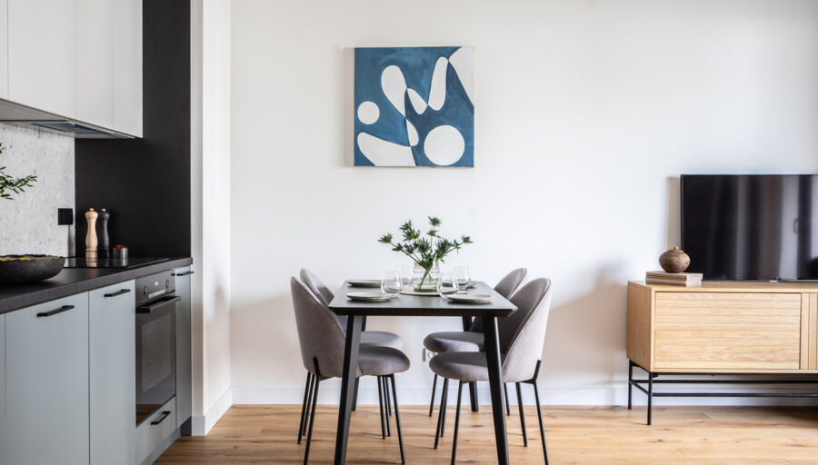 Nowoczesna jadalnia z czarnym stołem, czterema szarymi krzesłami i obrazem w odcieniach niebieskiego. W tle aneks kuchenny i drewniany stolik RTV.